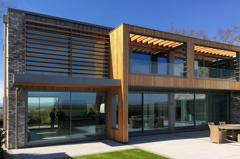 Modern two-story house with large glass windows, wooden paneling, and a patio with outdoor furniture.