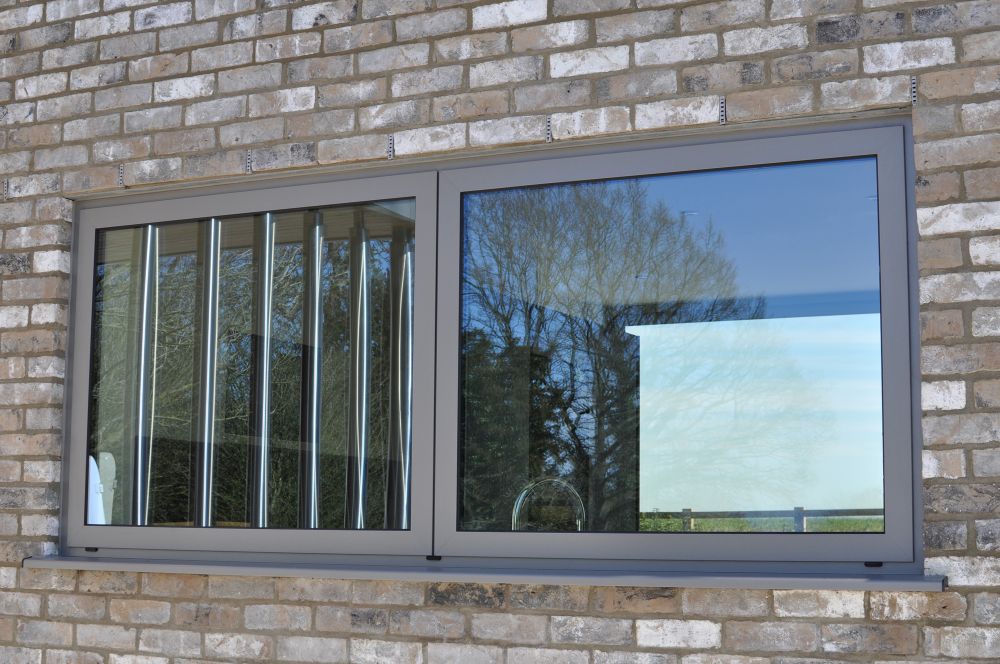 A modern window with a sliding pane is set in a brick wall. The reflection shows outdoor trees and sky.