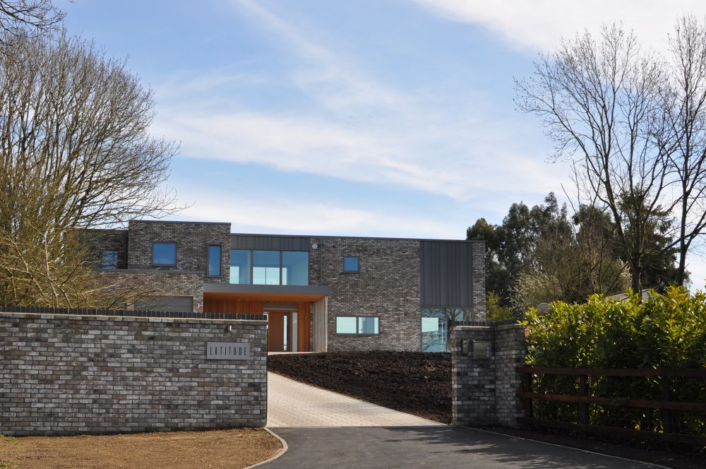 Modern brick house with large windows and a gated entrance, surrounded by trees.