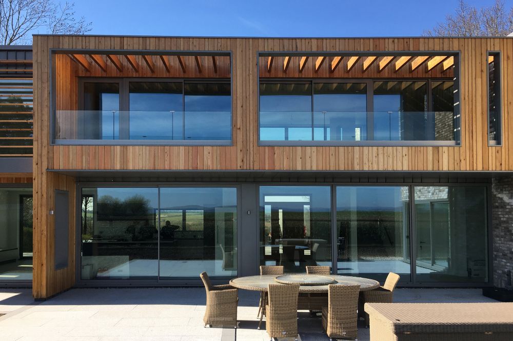 Modern two-story house with wooden paneling and large glass windows. A round outdoor table with chairs is set on the patio in front.