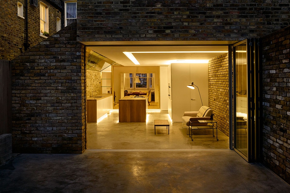 Brick-walled modern home with open sliding doors reveals a warmly lit interior featuring a kitchen, living area with a floor lamp, chair, and table, and a view of a cozy illuminated room beyond.