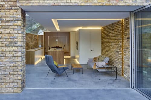 Modern living space with brick walls, featuring a lounge area with two chairs, a coffee table, and a dining area with a wooden table and chairs. Glass doors open to the outside.