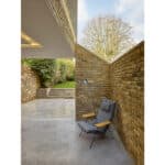 Thumbnail of http://Modern%20seating%20area%20with%20a%20gray%20armchair%20under%20a%20corner%20lamp.%20Exposed%20brick%20walls%20and%20concrete%20flooring.%20Glass%20doors%20open%20to%20an%20outdoor%20space%20with%20trees%20and%20a%20cloudy%20sky.