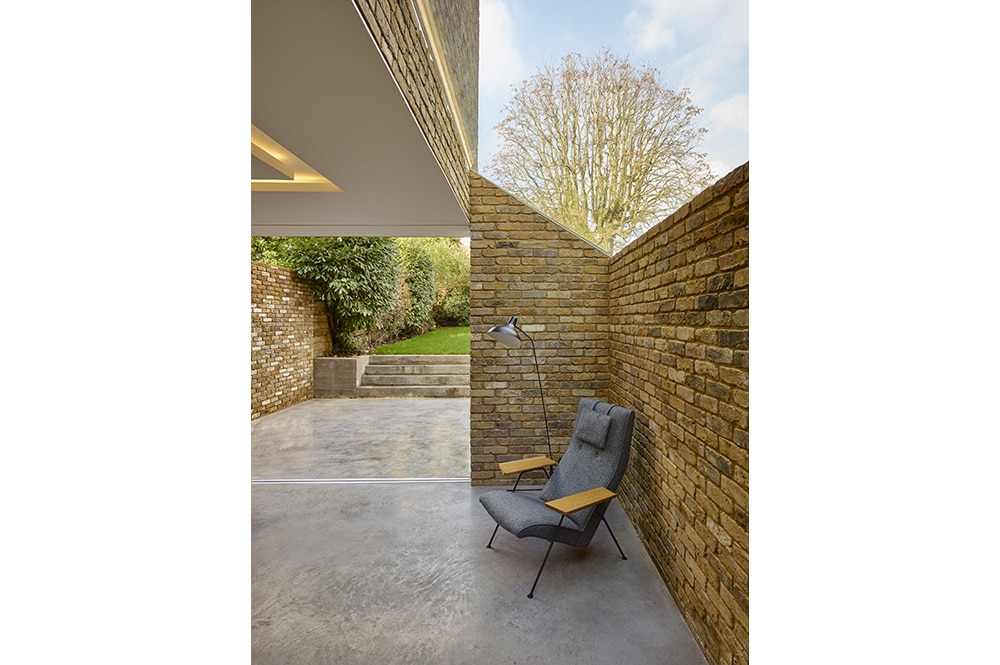 Modern seating area with a gray armchair under a corner lamp. Exposed brick walls and concrete flooring. Glass doors open to an outdoor space with trees and a cloudy sky.