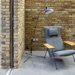Thumbnail of http://A%20modern%20gray%20lounge%20chair%20with%20wooden%20armrests%20is%20positioned%20next%20to%20a%20brick%20wall,%20underneath%20a%20black%20floor%20lamp,%20on%20a%20concrete%20floor.