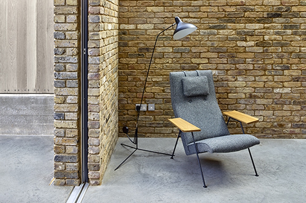 A modern gray lounge chair with wooden armrests is positioned next to a brick wall, underneath a black floor lamp, on a concrete floor.