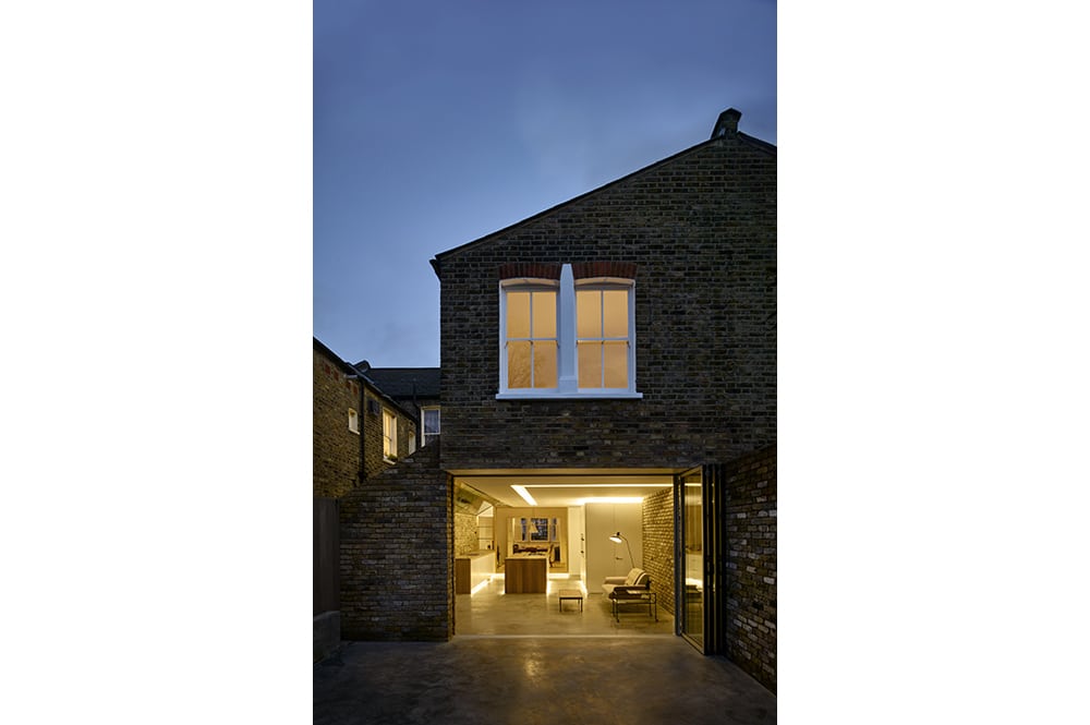 Exterior view of a two-story brick house at dusk, with illuminated interiors visible through large glass doors on the ground floor.