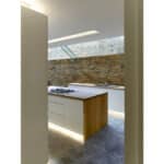 Thumbnail of http://Modern%20kitchen%20with%20white%20cabinetry,%20a%20wood-accented%20island%20with%20a%20stovetop,%20concrete%20floor,%20and%20exposed%20brick%20wall.%20Skylight%20provides%20natural%20lighting.