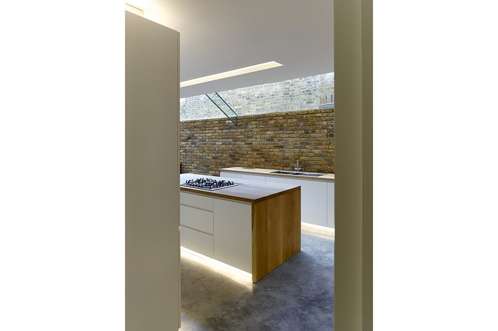 Modern kitchen with white cabinetry, a wood-accented island with a stovetop, concrete floor, and exposed brick wall. Skylight provides natural lighting.