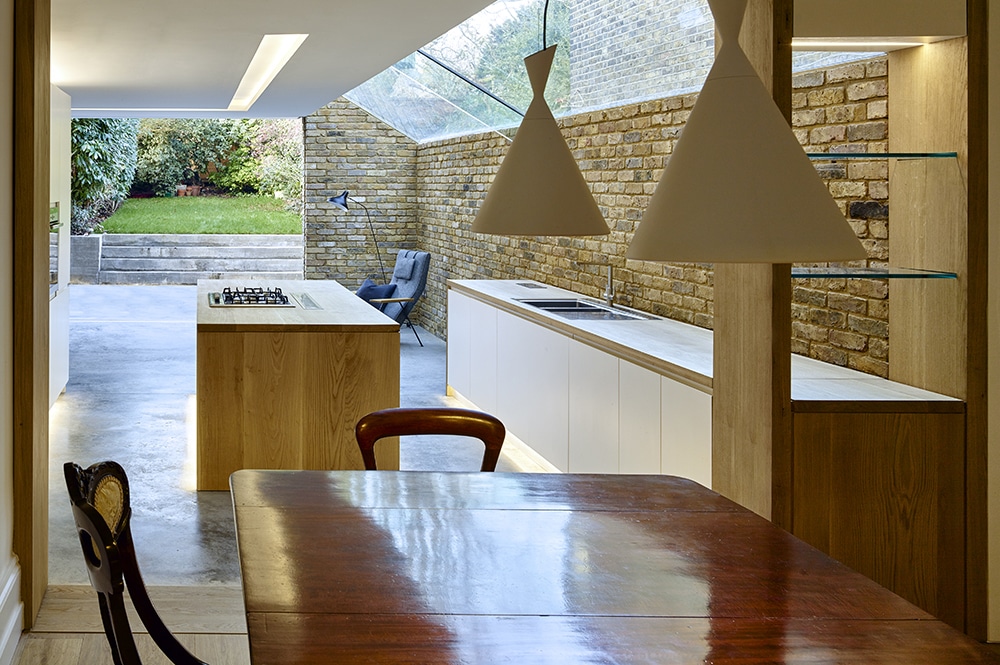 Modern kitchen with skylight, wood and brick accents, and garden view. Features an island with stovetop, white cabinets, and triangular hanging lights.