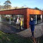 Thumbnail of http://Outside%20a%20modern,%20single-story%20house%20with%20large%20glass%20windows%20and%20a%20wooden%20exterior,%20three%20people%20stand%20in%20admiration%20of%20this%20architectural%20marvel%20straight%20out%20of%20Grand%20Designs.