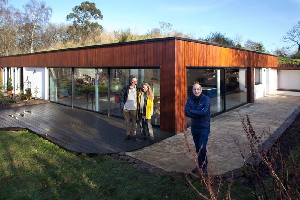 Outside a modern, single-story house with large glass windows and a wooden exterior, three people stand in admiration of this architectural marvel straight out of Grand Designs.