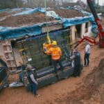 Thumbnail of http://Workers%20use%20a%20crane%20to%20lift%20a%20glass%20panel%20at%20a%20construction%20site%20reminiscent%20of%20Grand%20Designs,%20where%20the%20roof%20is%20innovatively%20covered%20in%20soil.%20A%20pickup%20truck%20is%20parked%20nearby,%20and%20trees%20are%20visible%20in%20the%20background.