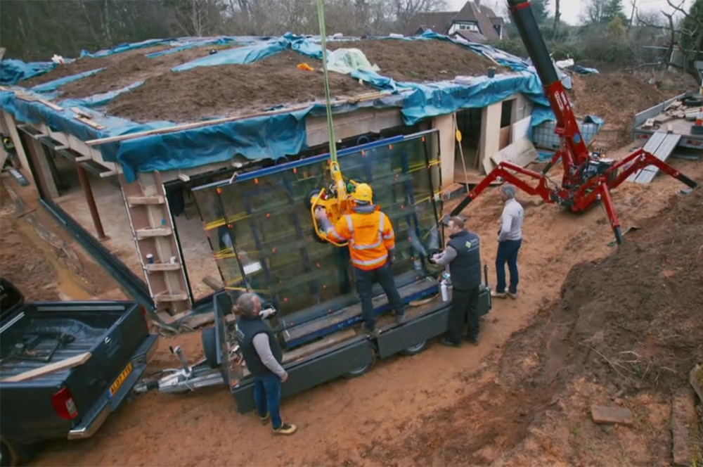 Workers use a crane to lift a glass panel at a construction site reminiscent of Grand Designs, where the roof is innovatively covered in soil. A pickup truck is parked nearby, and trees are visible in the background.