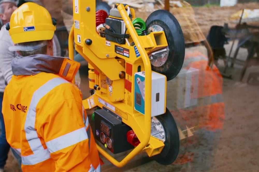 A worker in an orange safety jacket, reminiscent of "Grand Designs," skillfully operates a device with suction cups to lift a large glass panel at the construction site.