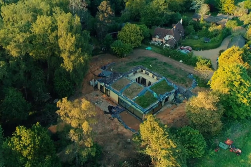 An aerial view reminiscent of a Grand Designs project shows a partially constructed house with a green roof nestled among trees, alongside another home.