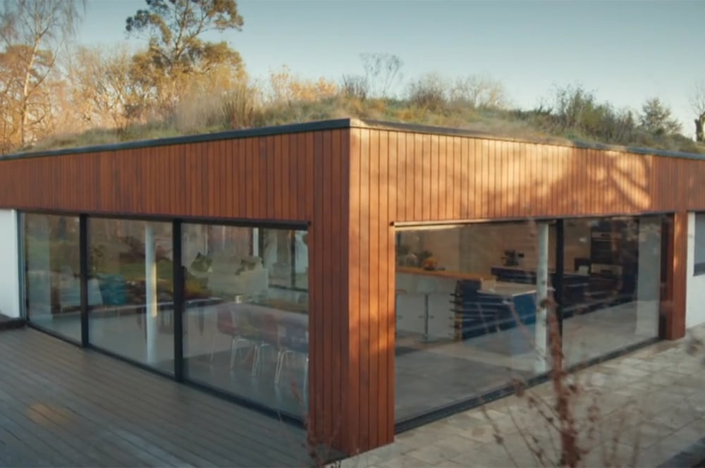 The Modern wooden house, reminiscent of a Grand Designs project, features large glass windows and a vibrant green roof. It is beautifully nestled among trees and grass under a clear sky.