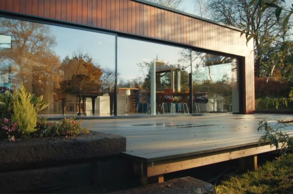 This modern building, reminiscent of a Grand Designs project, boasts large glass windows that reflect the trees and clear sky. In the foreground, a wooden deck seamlessly blends with the lush greenery surrounding the structure.