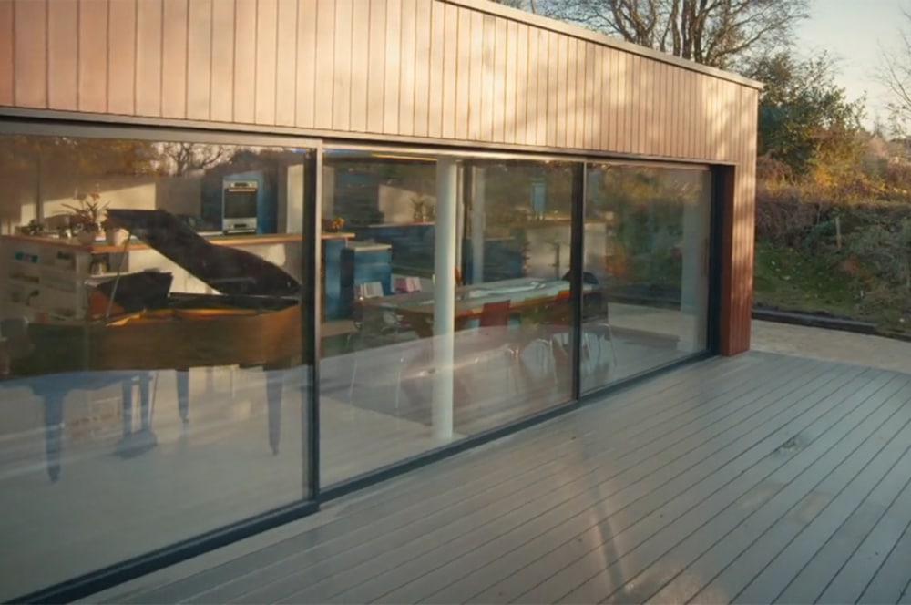 This modern house, reminiscent of Grand Designs, boasts large glass doors that unveil an interior featuring a grand piano and elegant furniture. The exterior is beautifully complemented by a wooden deck and lush surrounding greenery.