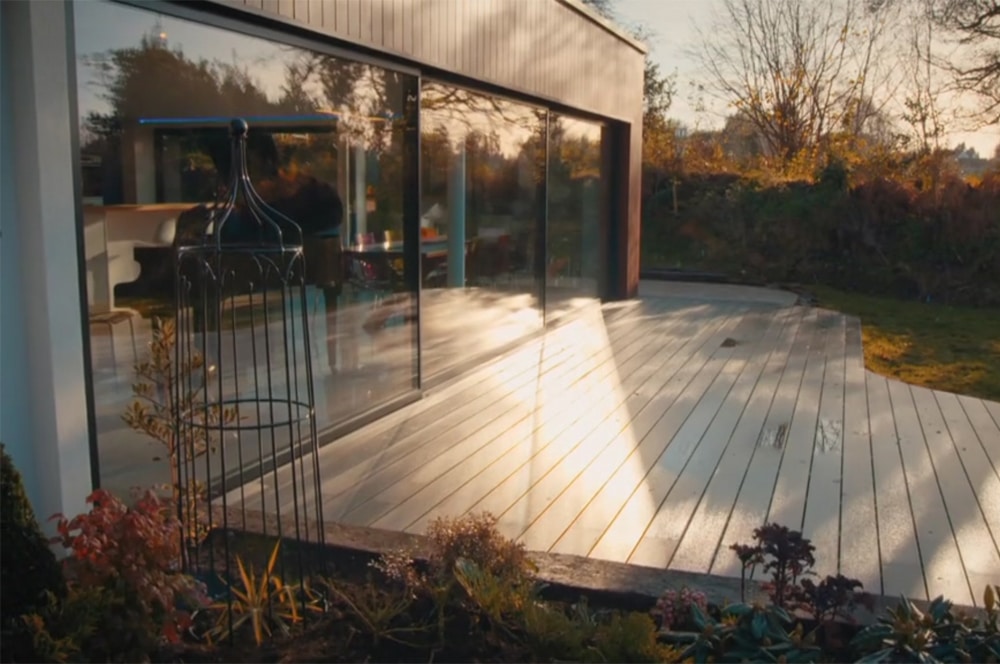 The modern house exterior, reminiscent of Grand Designs, features large glass sliding doors leading to a wooden deck nestled in a lush garden. Sunlight casts reflections on the deck, where an empty metal birdcage stands quietly to the left.