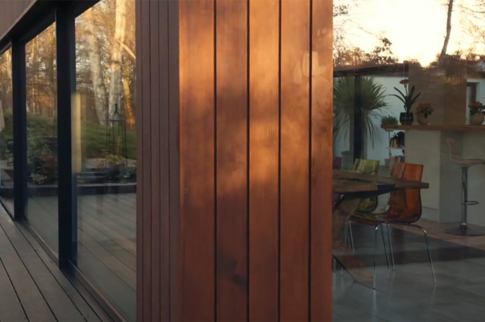 With echoes of Grand Designs, this modern house showcases large glass windows that reflect the surrounding trees, revealing an inviting dining area inside. The space features several colorful chairs and a bar counter adorned with lush plants, embodying both style and nature.