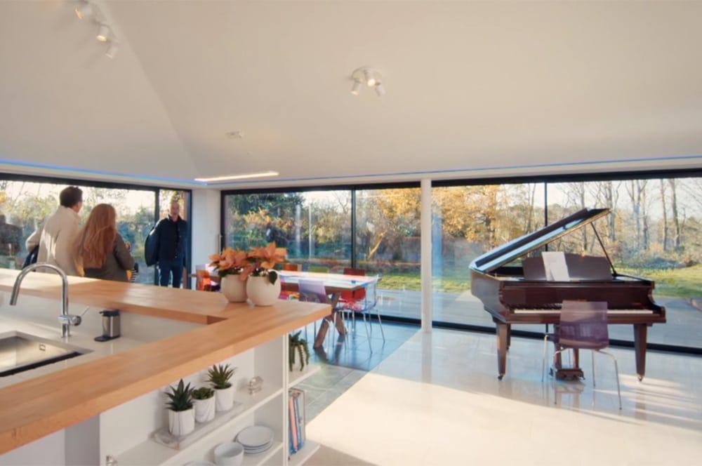 Spacious modern room with large windows, reminiscent of a Grand Designs masterpiece, featuring a grand piano and a kitchen island adorned with plants, while a group of people gathers near the entrance.
