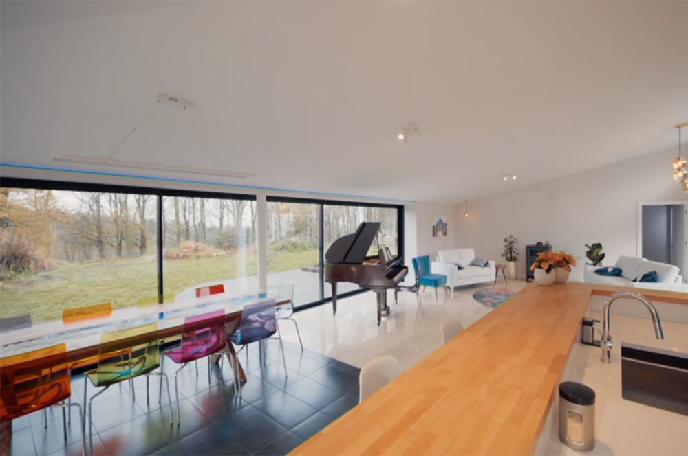Spacious modern living area reminiscent of Grand Designs, featuring a wooden counter, colorful chairs around a long table, a grand piano, and large glass doors opening to a garden view.