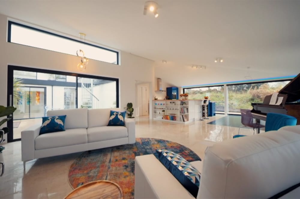 The spacious living room, reminiscent of Grand Designs, boasts white sofas, a round rug, and a piano. Expansive windows flood the space with natural light, while shelves brim with books and decor.