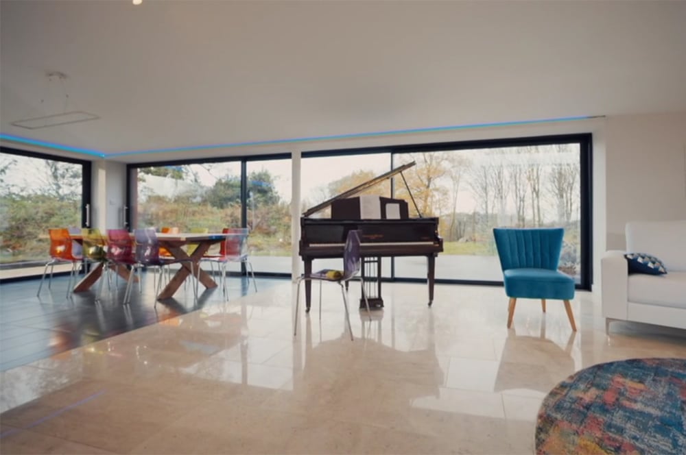 This Grand Designs-inspired spacious modern living room features a grand piano, large windows revealing a scenic view, and a dining area adorned with colorful chairs.