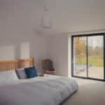 Thumbnail of http://A%20Grand%20Designs-inspired%20minimalist%20bedroom%20features%20a%20low%20wooden%20bed,%20white%20bedding,%20a%20chair,%20and%20a%20potted%20plant.%20Sunlight%20streams%20through%20large%20sliding%20glass%20doors%20overlooking%20the%20garden.