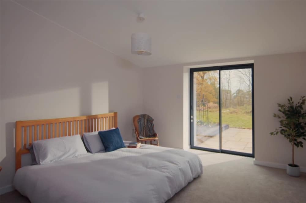 A Grand Designs-inspired minimalist bedroom features a low wooden bed, white bedding, a chair, and a potted plant. Sunlight streams through large sliding glass doors overlooking the garden.