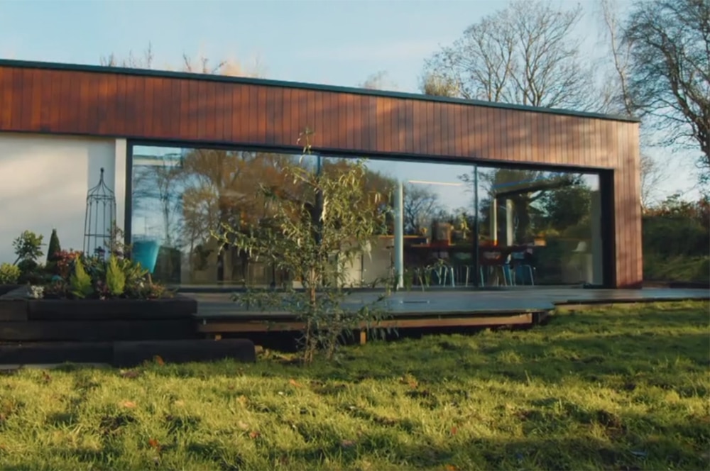 A modern one-story house with large glass windows, reminiscent of a Grand Designs feature, is nestled within a lush grassy yard and surrounded by trees.