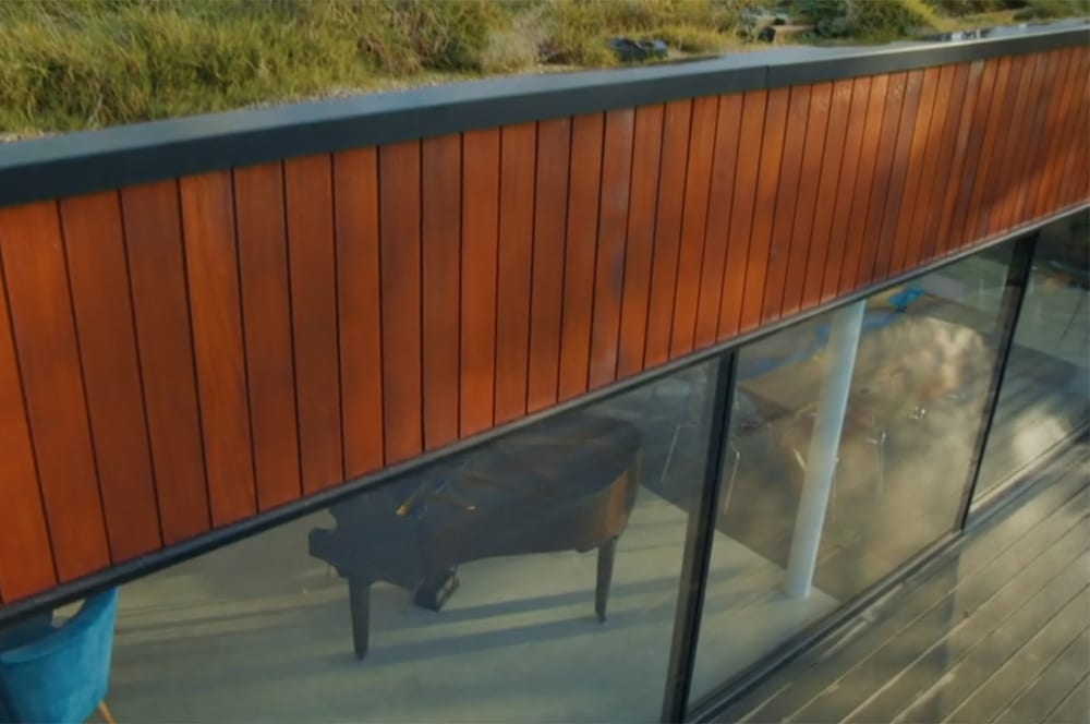 A grand piano is visible through the large windows of a modern Grand Designs building, boasting a wooden exterior and a grass roof.