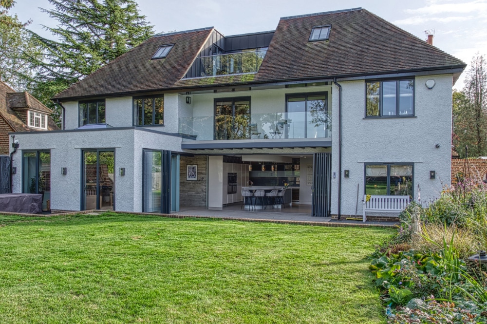 Modern two-story house with large windows, glass balconies, and an open patio leading to a well-kept lawn and garden in front.