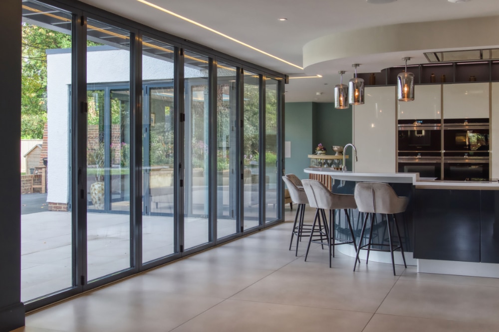Modern kitchen with a large island, bar stools, and pendant lights. Floor-to-ceiling glass doors open to a patio area. Neutral tones create a spacious and airy feel.