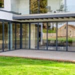 Thumbnail of http://Modern%20house%20with%20large%20glass%20folding%20doors%20opening%20to%20a%20grassy%20backyard,%20leading%20to%20a%20covered%20patio%20area%20under%20an%20upper%20balcony%20with%20glass%20railing.