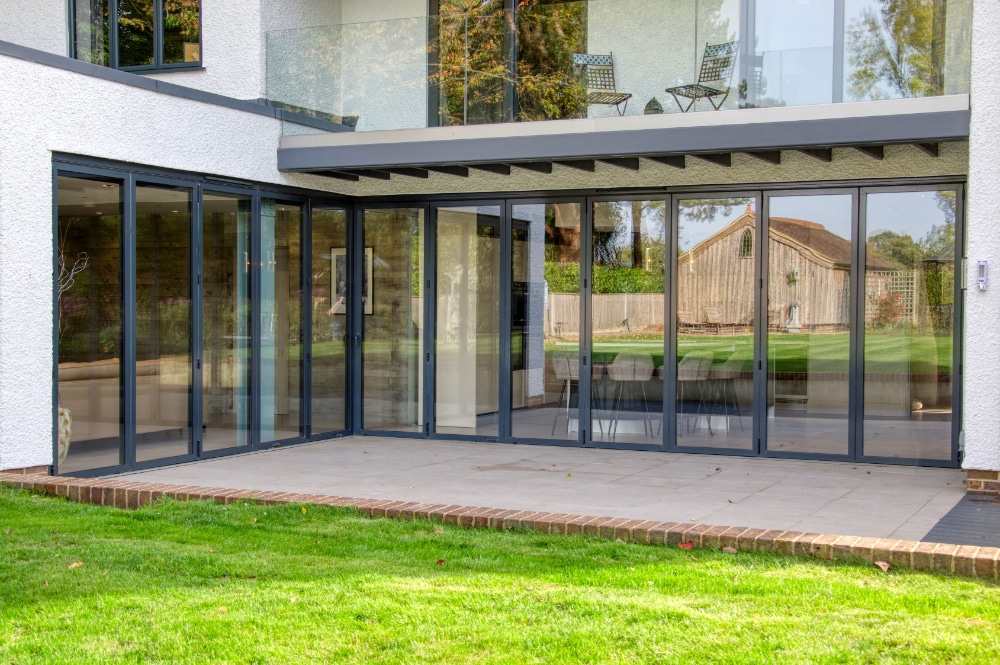 Modern house with large glass folding doors opening to a grassy backyard, leading to a covered patio area under an upper balcony with glass railing.