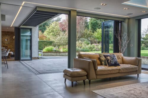 Spacious living room with a tan leather sofa, open sliding doors, and a view of a garden. Modern decor with a large rug and decorative branches in a vase.