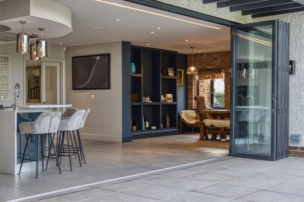 Modern open-concept living space with a kitchen island, bar stools, and a dining area. Sliding glass doors lead to a tiled patio. Dark shelving contrasts with light walls and flooring.
