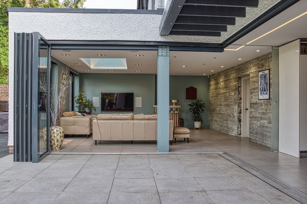 Modern open-plan living room with beige couches, wall-mounted TV, and glass folding doors leading to a patio. Walls are painted in green and gray with decorative plants and art.