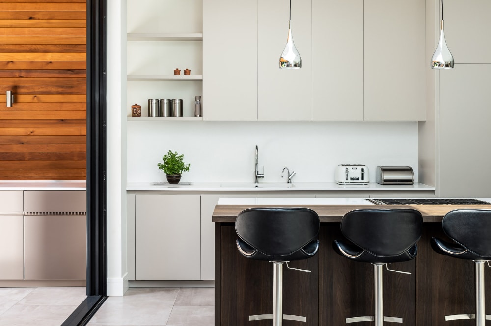 Modern kitchen with white cabinets, a sink, and countertop appliances. Three black bar stools at a dark wooden island. Hanging light fixtures and a potted plant add decor.