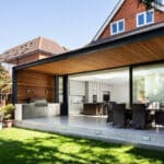 Thumbnail of http://Modern%20house%20exterior%20with%20a%20covered%20patio.%20Features%20an%20outdoor%20dining%20area%20and%20barbecue%20set.%20Large%20glass%20doors%20open%20to%20the%20kitchen%20and%20dining%20room.%20Brick%20walls%20and%20a%20green%20lawn%20are%20visible.