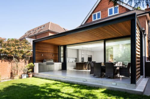 Modern house exterior with a covered patio. Features an outdoor dining area and barbecue set. Large glass doors open to the kitchen and dining room. Brick walls and a green lawn are visible.
