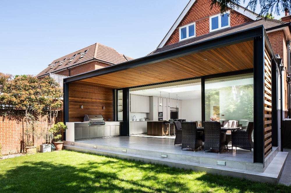 Modern house exterior with a covered patio. Features an outdoor dining area and barbecue set. Large glass doors open to the kitchen and dining room. Brick walls and a green lawn are visible.