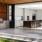 Thumbnail of http://Modern%20open-plan%20kitchen%20with%20a%20large%20island,%20barstools,%20and%20stainless%20steel%20appliances.%20Floor-to-ceiling%20sliding%20glass%20doors%20open%20out%20to%20a%20patio%20with%20outdoor%20dining%20furniture%20and%20a%20grill.