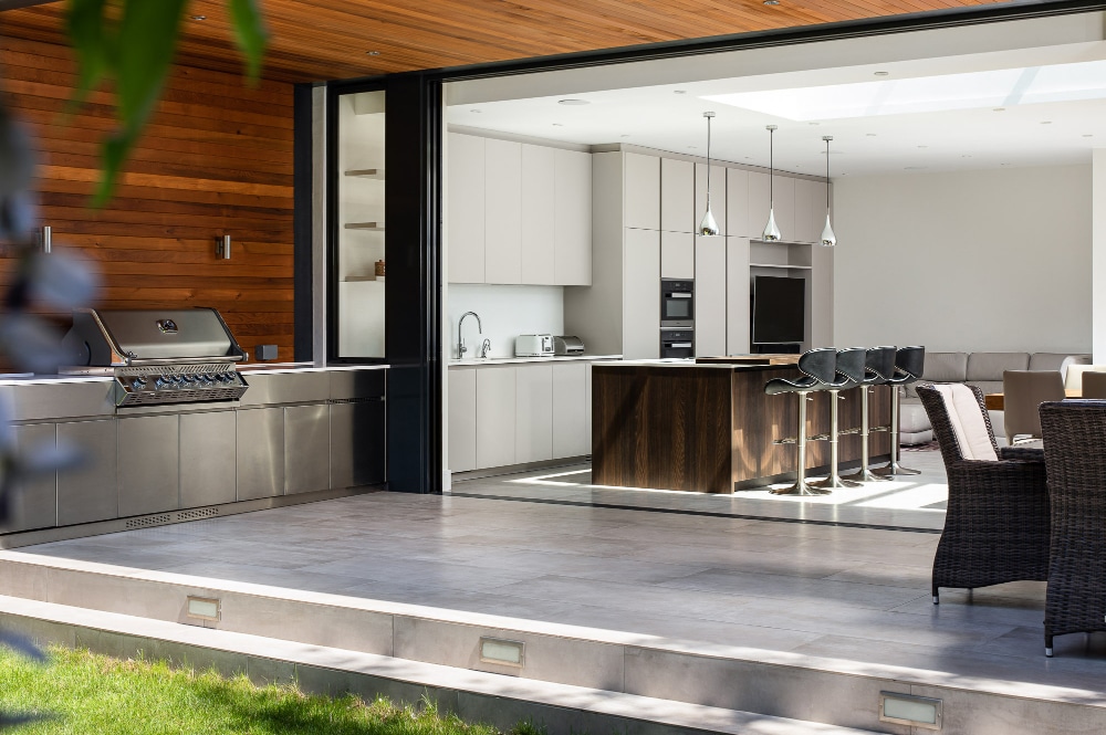 Modern open-plan kitchen with a large island, barstools, and stainless steel appliances. Floor-to-ceiling sliding glass doors open out to a patio with outdoor dining furniture and a grill.