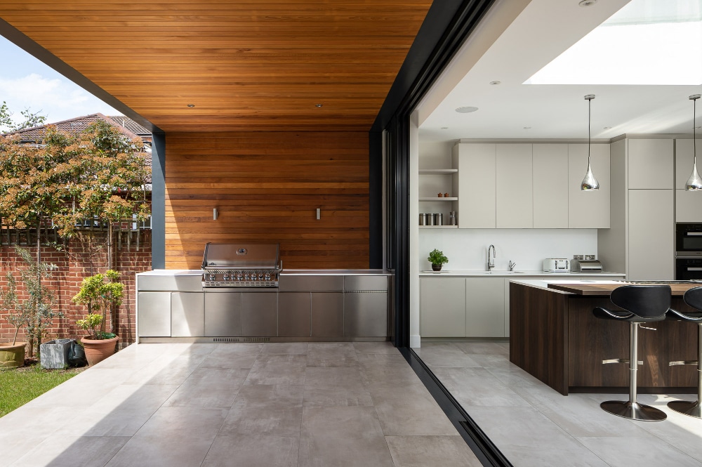 Modern kitchen with sliding glass doors, outdoor grill, wooden ceiling, and contemporary indoor fixtures. Features bar stools, potted plants, and ample natural light.