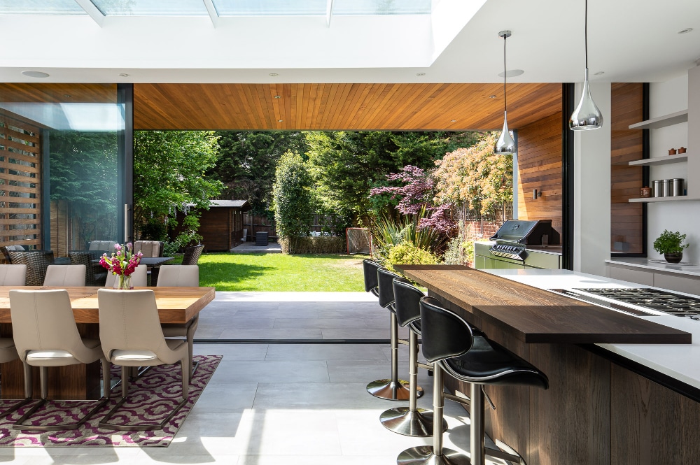 Modern kitchen with an open layout, featuring a bar counter and dining table. Glass doors lead to a garden with a grill and outdoor seating. Skylight and wood accents add natural warmth.