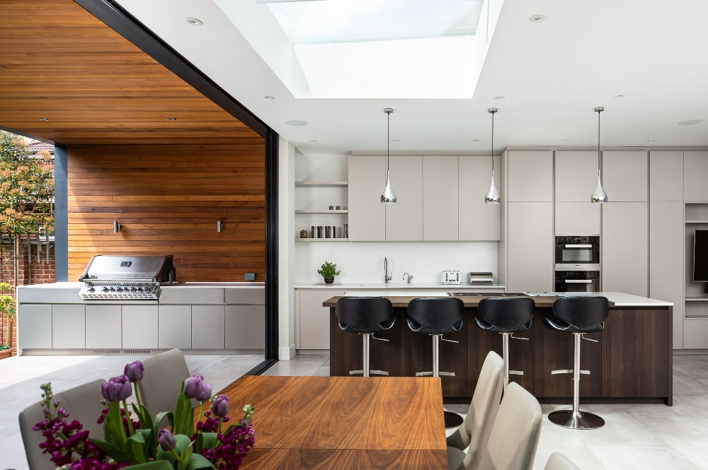 Modern kitchen with a skylight, a wooden dining table, bar stools at an island, built-in appliances, and an outdoor area with a grill.