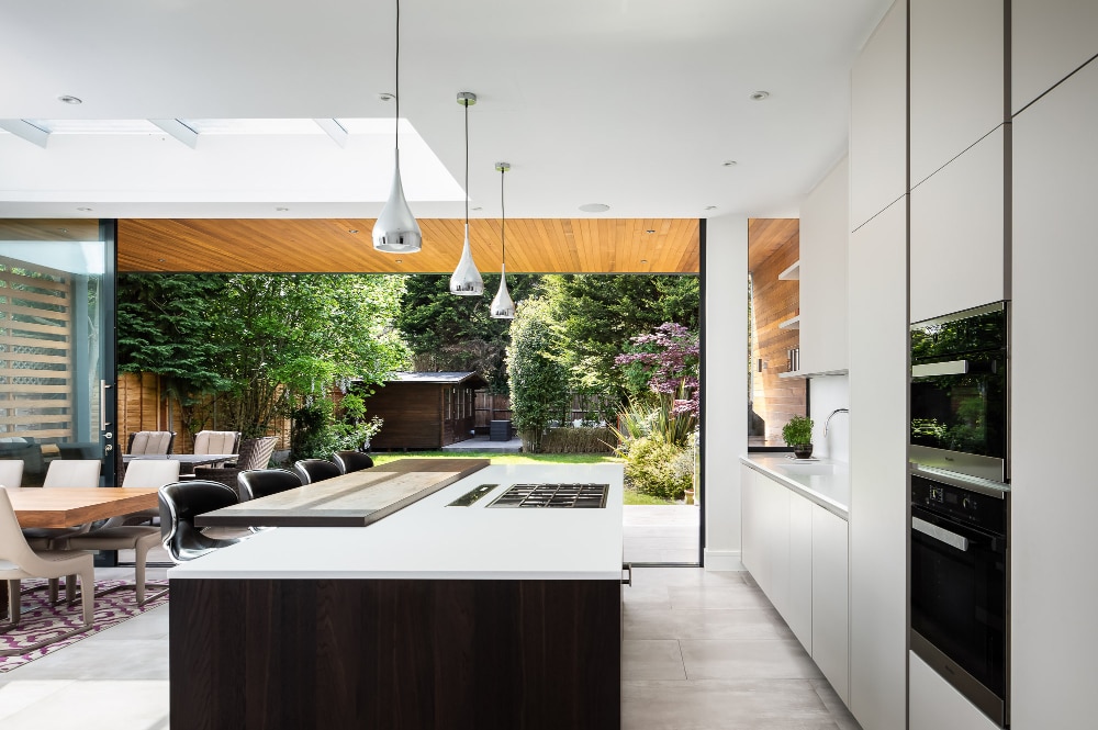 Modern kitchen with an island, pendant lights, and white cabinetry. Large sliding doors open to a garden with trees and greenery. Dining table and chairs are visible on the left.